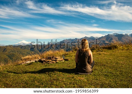 A blonde girl, sits on a plateau near the fireplace and looks at the endless mountain range and Mount Elbrus.
Blue cloudy sky over hills, green forest and alpine meadows.