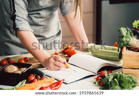 Unrecognizable woman is looking for recipes in cookbook.
Female chef reading recipes in book and holding pen on table full with fresh vegetables for healthy lunch Royalty-Free Stock Photo #1959865639