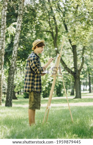 a young guy in a straw hat sits in the park in front of an easel and paints a picture with oil paints. Artistic work in nature. Plein air.