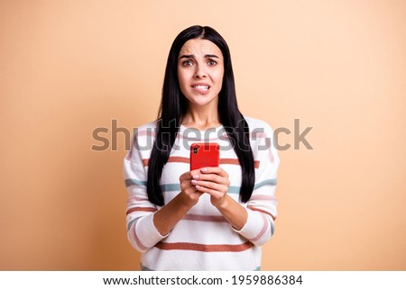Photo of young beautiful worried scared girl with long brown hair biting lips hold smartphone isolated on beige color background