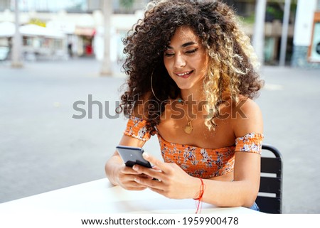 Beautiful colombian woman with afro hairstyle using mobile phone, flirting with someone or posting new photo on social media. Outdoor photo. Relax time in cafe. 