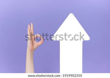 Unrecognizable young male makes thumb up gesture near white paper arrow, demonstrates approval or agreement, isolated on purple studio background. Body language concept. Hand sign. Like gesture