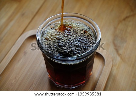Pouring freshly brewed black filter coffee into glass cup, close-up. Wooden table