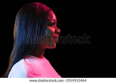 Calm, cheerful. African young woman's portrait on dark studio background in neon light. Beautiful female model in casual style. Concept of human emotions, facial expression, youth, sales, ad.