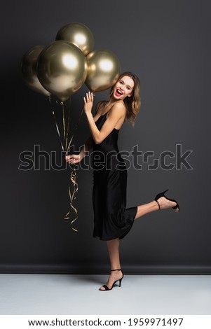 full length of amazed young woman in black slip dress holding golden balloons on grey