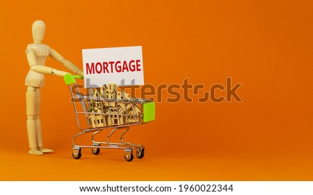 Mortgage symbol. Miniature shopping cart with wooden houses, the word mortgage. Wooden model of human. Beautiful orange background, copy space. Business and mortgage concept.