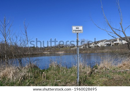 A sign in front of a lake that reads: “No fishing.” There are houses in the background. The sky is blue. Picture taken in St. Peters, Missouri.