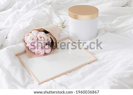 Gold gift box and white frame mockup on the bed. Bouquet of white peonies in craft packaging. Scandivanavian white interior.