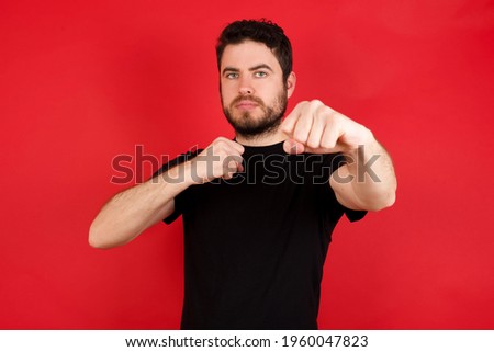 Young caucasian man wearing black t-shirt over red background Punching fist to fight, aggressive and angry attack, threat and violence