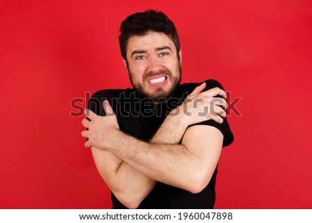 Desperate Young caucasian man wearing black t-shirt over red background trembles and feels cold, hugs oneself to warm up or feels scared notices something terrifying.