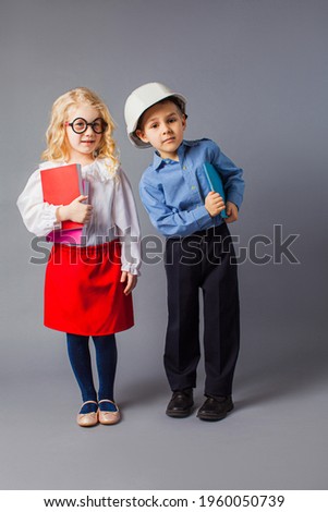 Two small children in the costumes of a teacher and an engineer at the gray background. The happy kids are holding books and looking at the camera