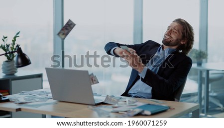 Young excited rich businessman employee sitting in office celebrating payday financial holiday throwing money rain. Emotions. Business success.