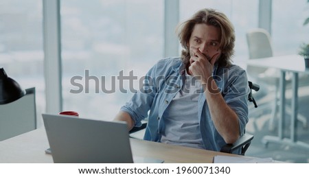 Frustrated young hesitating man using computer laptop sitting by desk doubting what to do. Self-doubting disabled man in wheelchair working first day at office. Royalty-Free Stock Photo #1960071340