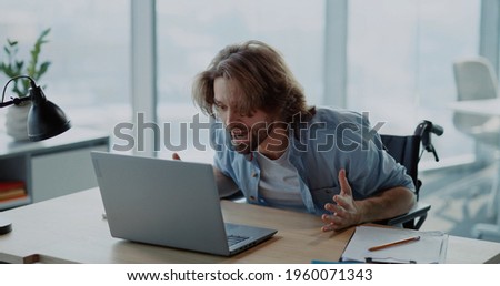 Frustrated young hesitating man using computer laptop sitting by desk doubting what to do. Self-doubting disabled man in wheelchair working first day at office. Royalty-Free Stock Photo #1960071343