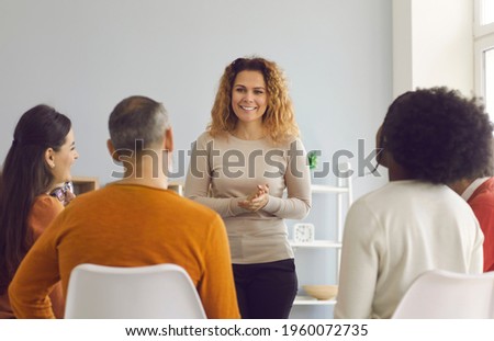 Happy beautiful woman making presentation standing in front of diverse audience. Psychologist or coach talking to group of young and mature people. Team of company workers listening to female speaker Royalty-Free Stock Photo #1960072735