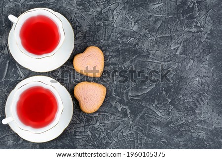 Two cups of raspberry tea on saucers and two beautiful heart-shaped cookies on a black background. Space for the text. Valentine's day, birthday, anniversary. Romantic breakfast.