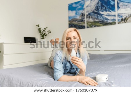 Sick beautiful female in blue cloth hold nasal mask with respiratory problem in hospital room. woman patient inhalation therapy by the mask of inhaler with soft stream smoke from bronchodilator. Royalty-Free Stock Photo #1960107379