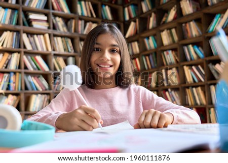 Happy smiling indian latin girl sitting at table learning at home classroom, having online video call. Portrait of adorable cute Mexican teen student child looking at camera, webcam view.