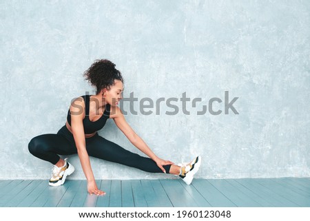 Portrait of fitness smiling black woman in sports clothing with afro curls hairstyle.She wearing sportswear.Young beautiful model stretching out before training.Female sitting in studio near gray wall Royalty-Free Stock Photo #1960123048