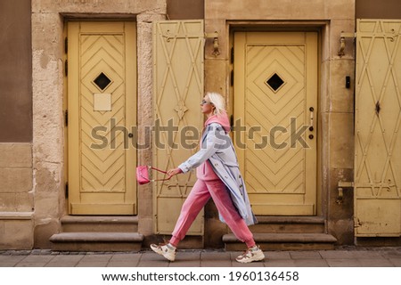Street style, fashion: woman wearing  trendy sport chic outfit walking in street of European city. Blue trench coat, sunglasses, pink hoodie, trousers, sneakers, mini bag. Copy, empty space for text Royalty-Free Stock Photo #1960136458