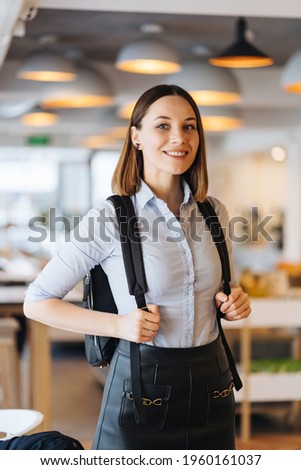 Indoor photo of young European Caucasian woman isolated dressed in blue shirt with backpack smiling happilyin a shop, restaurant, office