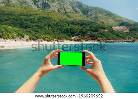 Man's hands hold a smartphone with a blank screen horizontally against a background of greenery and blue sea