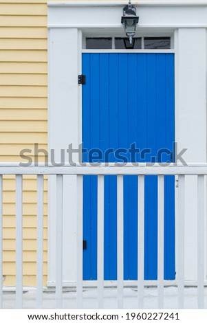 A bright royal blue door with white trim and a small window in a yellow building covered in clapboard. There's a small light over the exterior door. There's a white wooden handrail in the foreground.