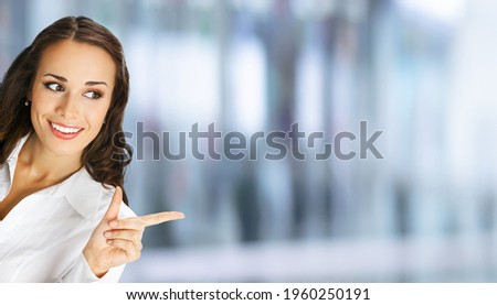 Young happy smiling woman in white confident blouse, showing pointing advertising at copy space. Business ad concept. Blurred modern office interior background. Brunette businesswoman, indoors. 