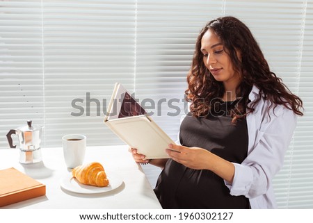 Side view of pregnant woman reading book while having morning breakfast with coffee and croissants on background of blinds. Good morning concept and pleasant lunch break