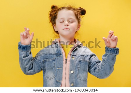 Dreamful small cute kid with eyes closed praying, wishing good luck or miracle. Superstitious little preschool girl crossing fingers, hoping her wish come true, standing isolated on yellow background