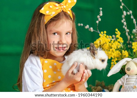 Easter closeup portrait of kid girl with rabbit on green background. Child farmer in yellow apron smiling, holding small white fluffy bunny. Alice in wonderland. Rural animal. Allergy. Happy childhood