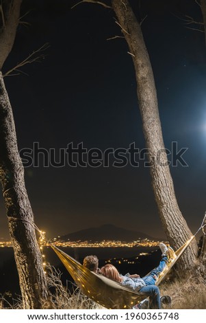 Couple relaxing in a hammock and enjoying starry sky. View from the top of the mountain to the lights of the city of Naples. Volcano Vesuvio. Sea and romance. Vertical photo. Sorrento Italy