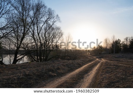 Rural dirt road in a field in early spring in the rays of the rising sun against the background of bare trees and the river. Spring landscape