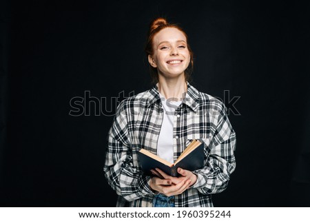 Laughing young woman college student holding opened book and looking at camera on black isolated background. Pretty redhead lady model emotionally showing facial expressions in studio, copyspace.