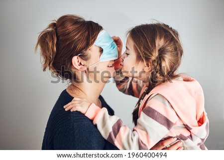 Little girl moving her mom's face mask on eyes and giving a kiss her mother. Funny moments during quarantine and covid-19 pandemic