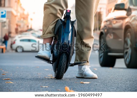 The legs of a young woman ready to ride an electric unicycle, abbreviated EUC, against the background of evening or morning city traffic. An unrecognizable woman on an electric monowheel.