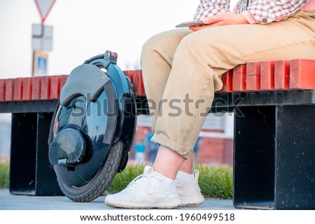 An unrecognizable young woman sitting on a park bench with a smartphone in her hands next to an electric unicycle, abbreviated EUC. The concept of using an electric mono wheel for daily trips. Royalty-Free Stock Photo #1960499518