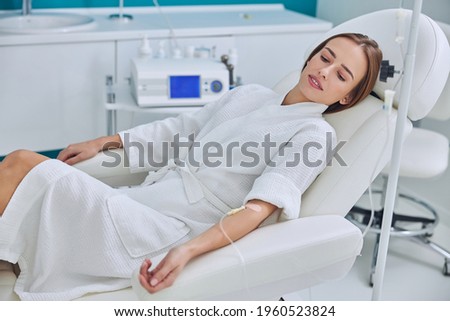 Redheaded beautiful female resting and getting IV infusion in spa salon Royalty-Free Stock Photo #1960523824