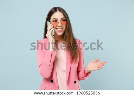 Young excited smiling woman 20s in pastel pink clothes glasses talking on mobile phone conducting pleasant conversation isolated on blue background studio portrait People lifestyle technology concept