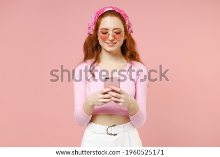 Young fun smiling happy caucasian woman 20s wear rose clothes bandana glasses using mobile cell phone chat online browsing isolated on pastel pink background studio portrait. People lifestyle concept