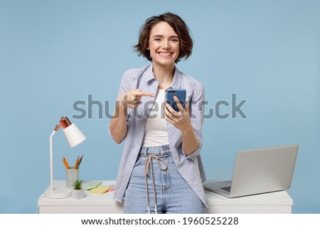 Young happy fun secretary employee business woman 20s in casual shirt work stand at white office desk with pc laptop point index finger on mobile cell phone isolated on pastel blue background studio.