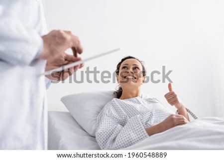 happy african american woman showing thumb up near doctor with digital tablet on blurred foreground