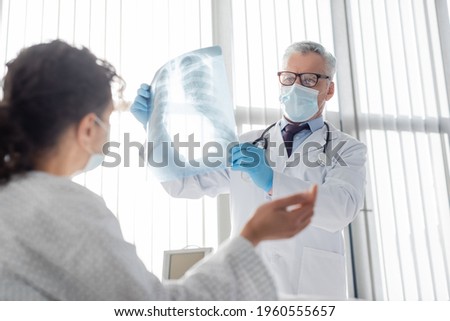 radiologist in medical mask holding lungs x-ray near african american woman on blurred foreground Royalty-Free Stock Photo #1960555657
