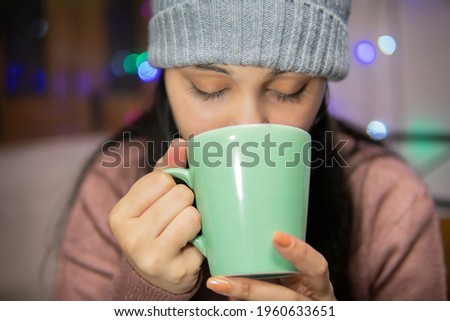 Indoor image of an Asian, Indian beautiful, serene young woman drinking coffee winter season at home. She is wearing warm clothes, sweater and a woolen cap.