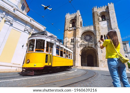 Young woman tourist photographing with phone famous retro yellow tram 28 on the street in Lisbon city in front of Santa Maria cathedral, Portugal.