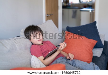 Authentic Kid sitting on sofa watching cartoons or playing games on tablet,Child boy using digital pad learning lesson on internet,Home schooling,Distance learning online education concept