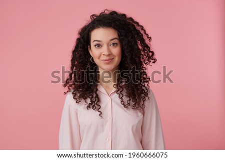 Charming girl, happy looking woman with dark long hair. Wearing earrings and pastel pink shirt. Has make up. People and emotion concept. Watching at the camera isolated over pastel pink background
