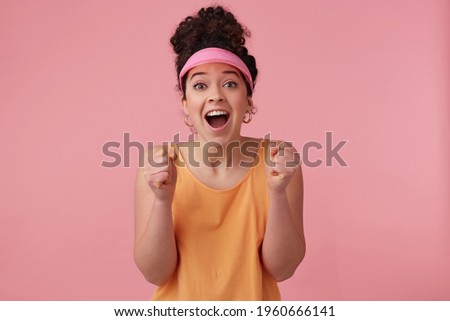 Cheerful woman, positive girl with dark curly hair bun. Wearing pink visor, earrings and orange tank top. Has make up. Clench fists in excitement. Watching at the camera isolated over pink background