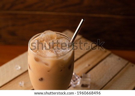 In a glass filled to the top with ice and coffee with milk. A glass of iced coffee sits on a crate against a dark wooden background.