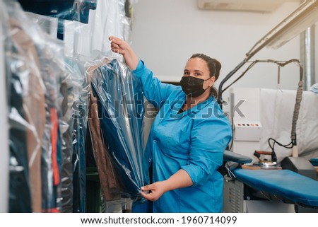 Woman in her 30s looking at the camera and placing the suit jacket that she has ironed on a hanger where she keeps the other clothes until the clients pick them up Royalty-Free Stock Photo #1960714099